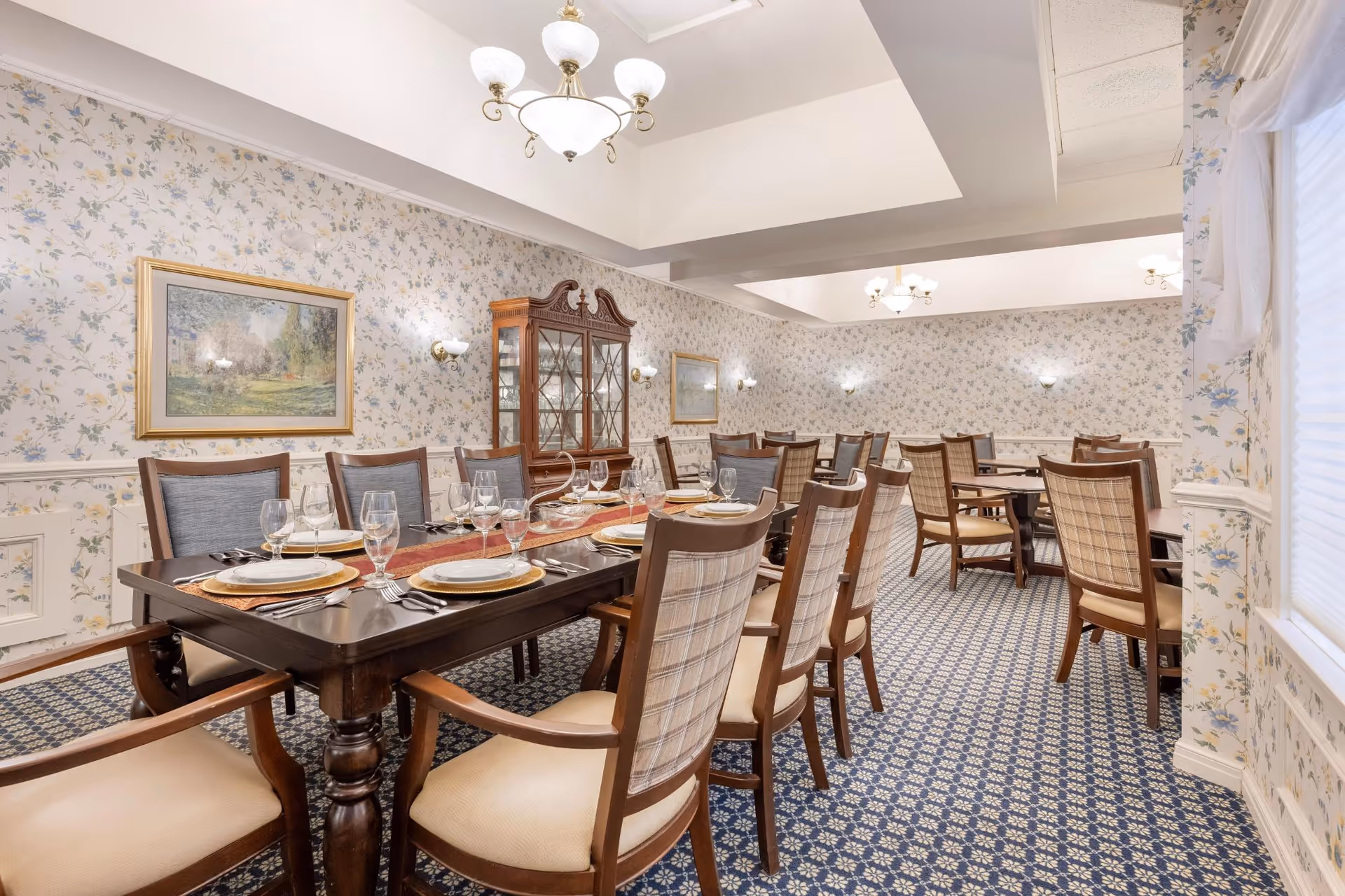 Formal dining room with a long set table, upholstered wooden chairs, a china cabinet, floral wallpaper, and chandeliers.