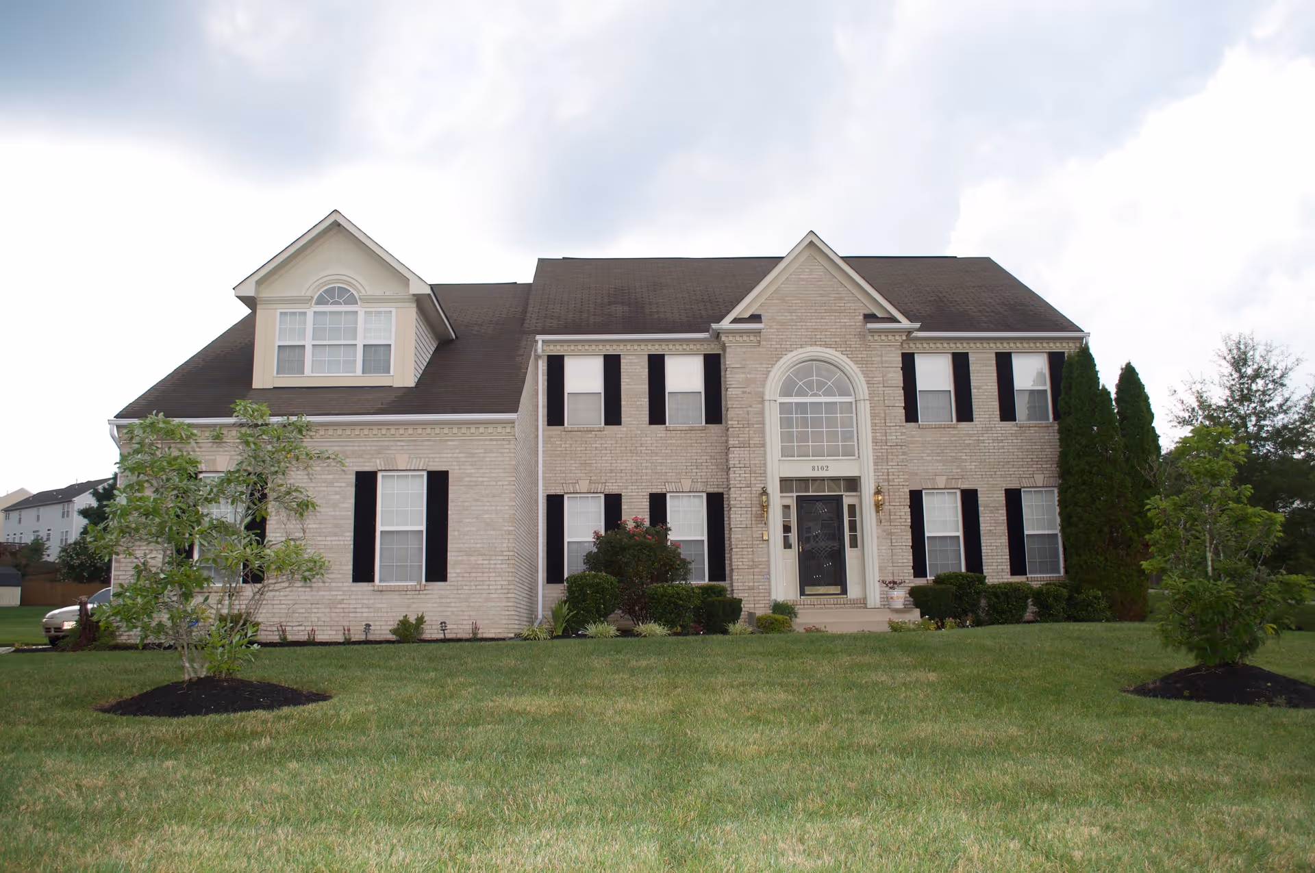 Front exterior view of a large two-story brick house with black shutters, a dark roof, and a well-maintained lawn with small trees and shrubs.
