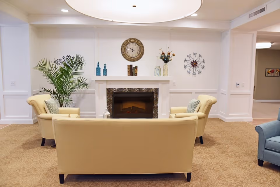 A cozy sitting area with a beige sofa and two matching armchairs arranged around a fireplace. The fireplace has a mosaic tile surround and a white mantel decorated with vases, books, and flowers. Above the mantel is a round clock. The walls are white with decorative molding, and there is a large green plant to the left. The carpet is light brown, and there is a blue armchair partially visible on the right.