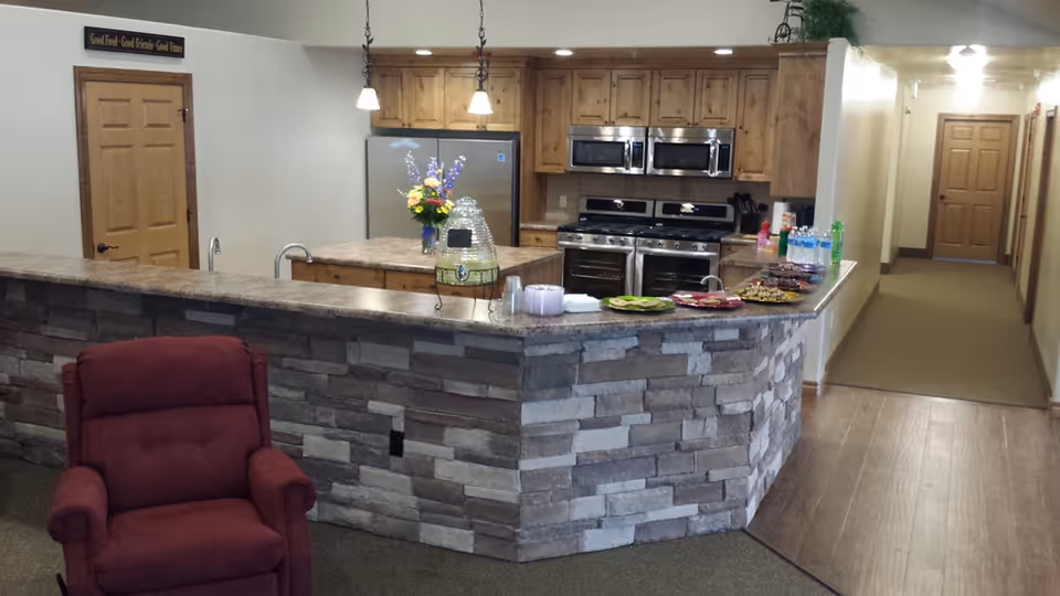 Interior view of a senior living facility kitchen area with a stone-faced counter and a maroon recliner chair in the foreground. The kitchen features wooden cabinets, stainless steel appliances including two ovens and two microwaves, and a refrigerator. On the counter, there are plates with snacks, bottled water, and a beverage dispenser. A hallway with wooden doors is visible to the right.
