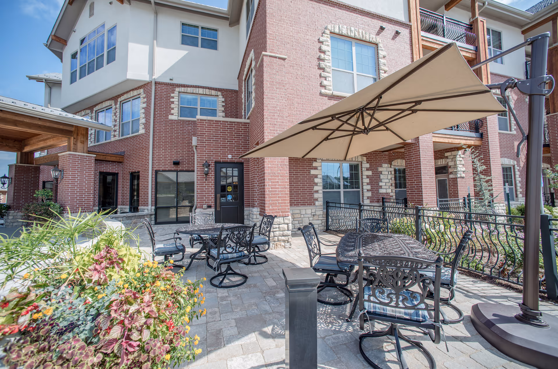 Outdoor patio area at The Village of Bedford Walk featuring metal tables and chairs with decorative designs, a large beige umbrella providing shade, and a brick building with multiple windows in the background. There are plants and flowers in the foreground adding greenery to the space.