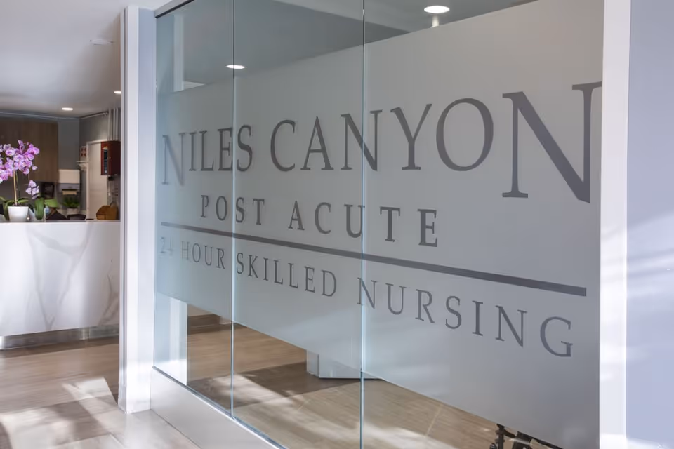 Interior view of Niles Canyon Post Acute facility showing a frosted glass wall with the facility name and '24 Hour Skilled Nursing' text. A reception desk with a purple orchid plant is visible in the background.