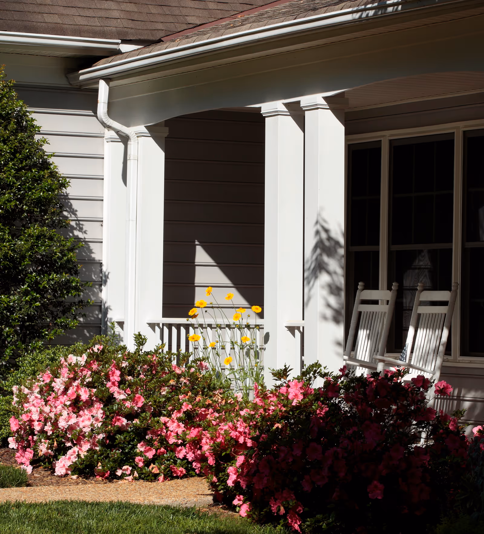 Front porch of a house with white pillars, two white rocking chairs, and a garden bed filled with blooming pink and yellow flowers in front.
