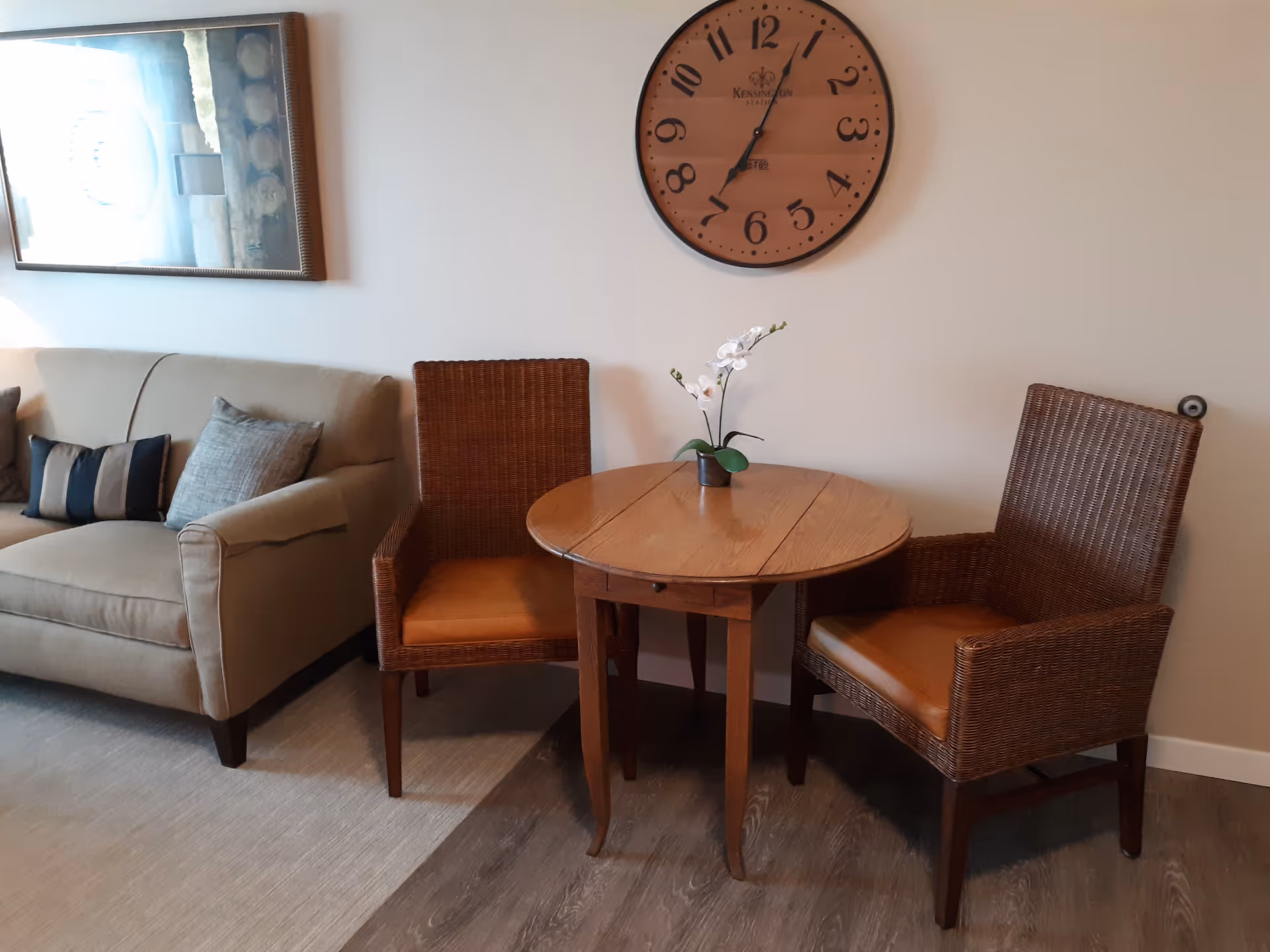 A cozy interior corner featuring a small round wooden table with a white orchid plant in a pot on top, flanked by two wicker chairs with brown cushions. To the left, there is a beige sofa with several decorative pillows. A large round wall clock and a framed artwork hang on the light-colored wall behind the furniture.