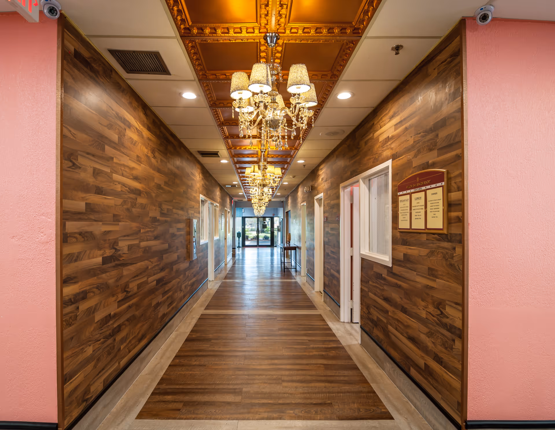 A long, well-lit hallway with wood-paneled walls and a wooden floor. The ceiling features ornate golden panels with multiple chandeliers hanging down the center. There are several doorways along the hallway and a glass exit door at the far end. The walls near the entrance are painted pink, and there is a sign with information mounted on the right wall.