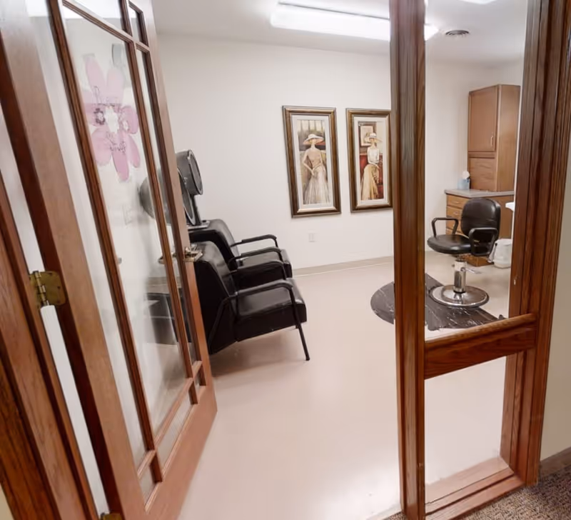 Interior view of a hair salon room in a senior living facility with wooden framed glass doors, two black salon chairs, a hair dryer station, wooden cabinets, and two framed paintings of women in vintage dresses on the wall.