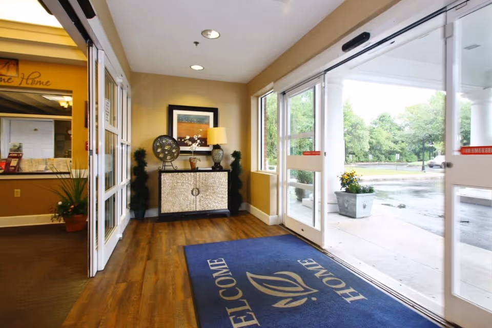 Entrance lobby with sliding glass doors open to the outside, a 'Welcome' mat on wood floors, and a decorative console table under a framed picture.