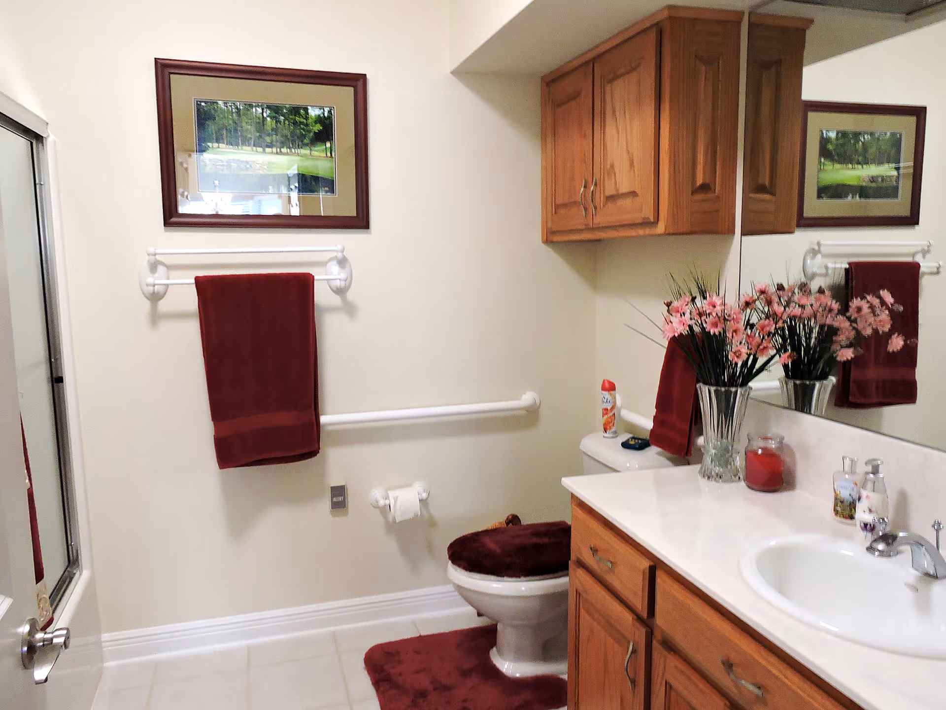 A clean bathroom with a white toilet, a white sink with wooden cabinets underneath, and a large mirror above the sink. The toilet has a burgundy cover and matching burgundy bath mat. There is a burgundy towel hanging on a white towel rack on the wall. A framed picture hangs above the towel rack. A vase with pink flowers, a red candle, and soap dispensers are on the countertop. The bathroom has white tiled flooring and a shower with a glass door on the left side.