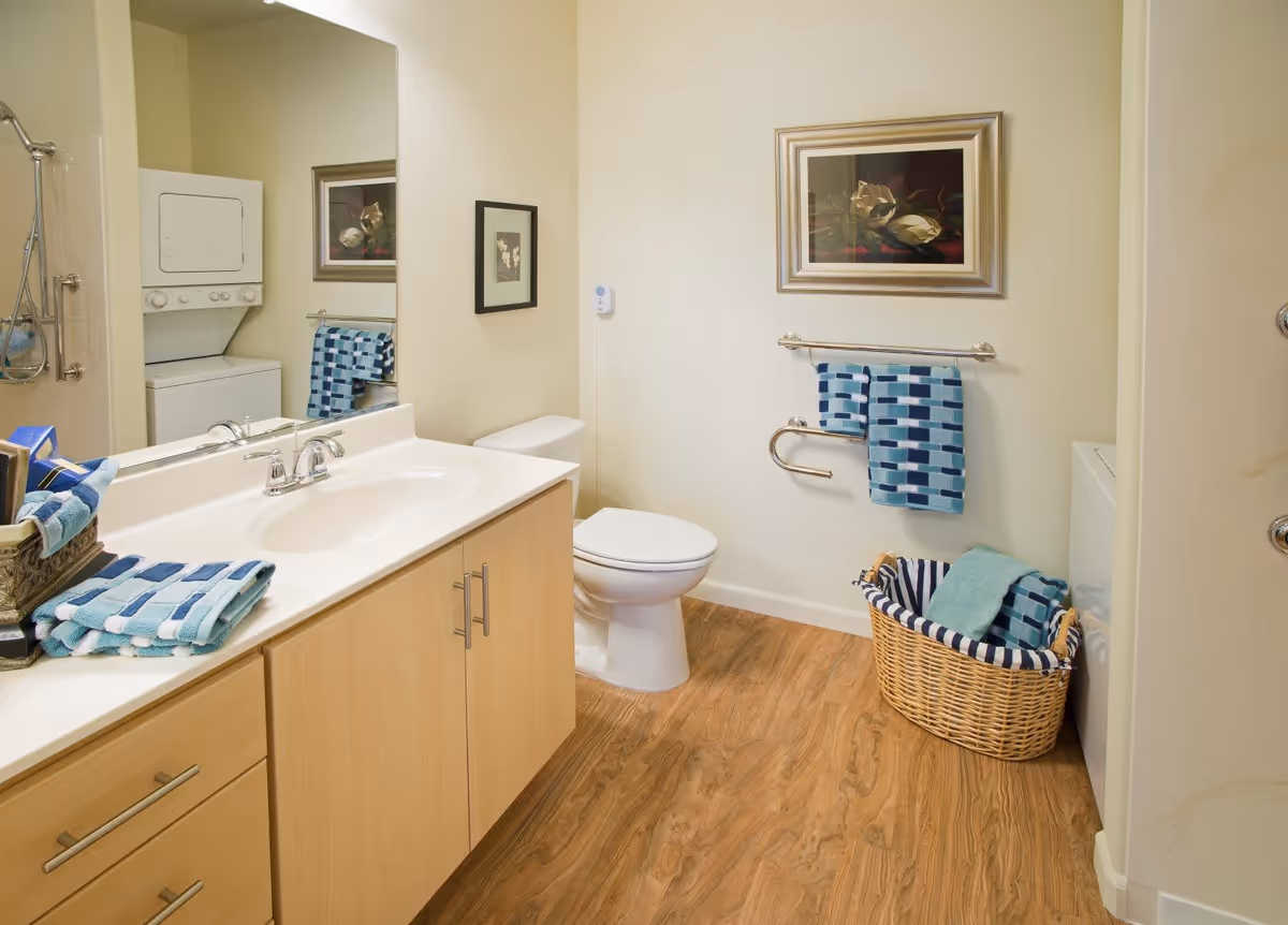 A clean bathroom with a wooden floor, a white sink with a wooden cabinet underneath, a toilet, and a shower area. There are blue and white patterned towels hanging on a towel rack and folded on the counter. A wicker basket with towels is placed on the floor next to a stacked washer and dryer unit. Two framed floral pictures hang on the walls.