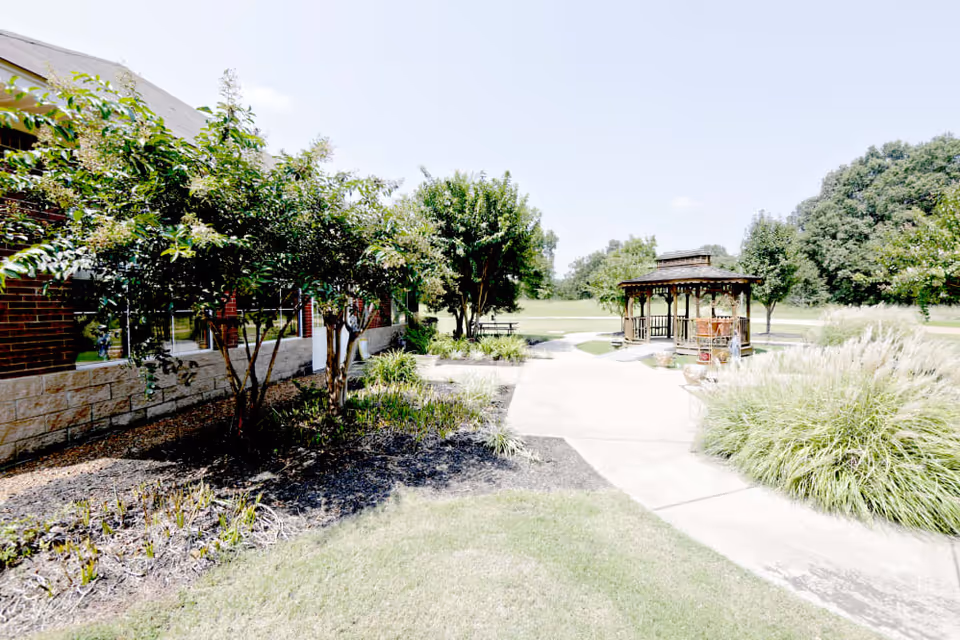 Outdoor garden area at Azalea Commons of Batesville featuring a paved walkway, green shrubs, trees, and a wooden gazebo under a clear sky.