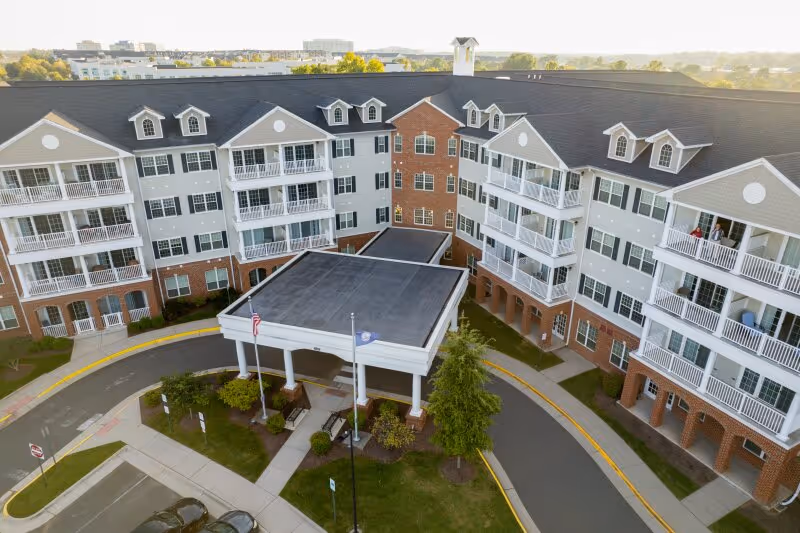 Aerial view of the front entrance of a multi-story senior living facility with balconies, a covered drop-off area, and landscaped greenery. The building has a combination of brick and light-colored siding with multiple windows and a circular driveway.