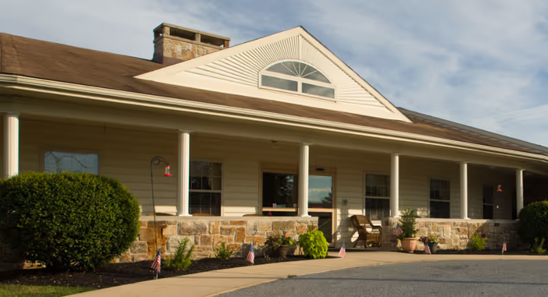 Exterior view of a single-story building with beige siding and stone accents, featuring a covered porch with white columns, potted plants, small American flags, and a wicker chair near the entrance under a partly cloudy sky.