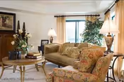 A cozy living room with a beige sofa, floral patterned armchair, round wooden coffee table with books and decorative items, two table lamps, a large window with curtains, and a potted plant in the corner.