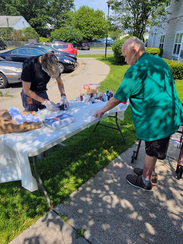 Two people outdoors at Albany Center Adult Home, one person standing and working on a table covered with a white cloth and various bottles, while the other person with a walker watches. They are on a sidewalk next to a grassy area with parked cars and a building in the background.