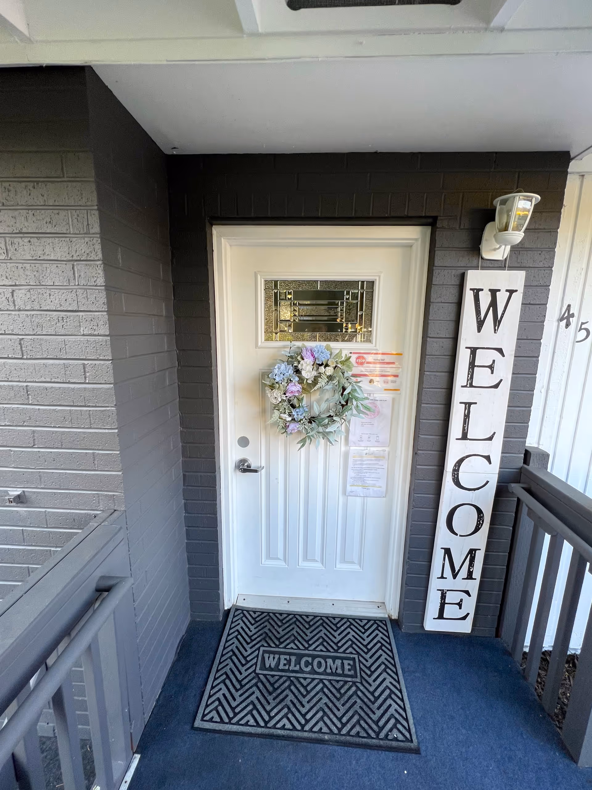 White front door set in a gray brick entryway with a floral wreath, a welcome mat, and a vertical 'WELCOME' sign.