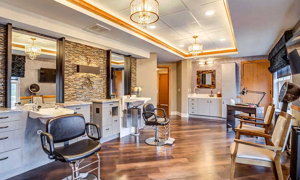 Interior of a stylish salon area with two black salon chairs in front of sinks and mirrors, wood flooring, modern lighting fixtures on the ceiling, and additional seating with wooden chairs near a window with black blinds.