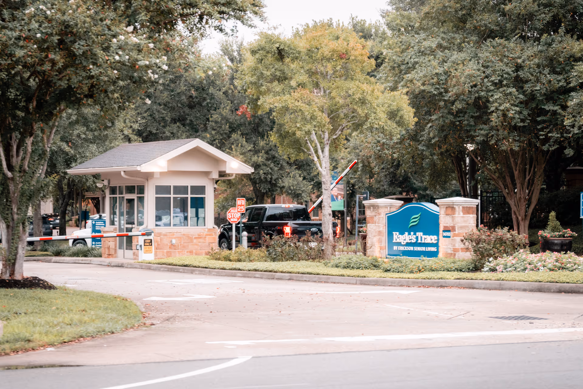 Entrance gate to Eagle's Trace senior living facility with a guardhouse, a black pickup truck passing through, surrounded by trees and landscaping.