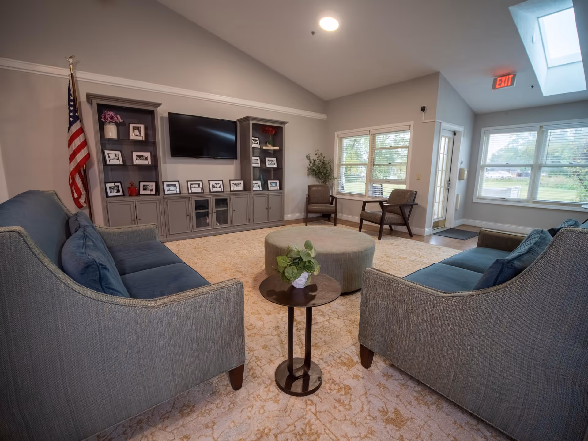 A cozy living room area with two gray sofas facing each other, a round ottoman in the center, and a small round side table with a potted plant. There is a large built-in entertainment center with a flat-screen TV and framed photos. Two armchairs are placed near windows that let in natural light. An American flag stands in the corner, and an exit sign is visible near the door.