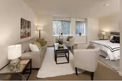 A bright and spacious living area in a senior living facility featuring a beige sofa with cushions, two beige armchairs, a glass coffee table on a white rug, a bed with white and black bedding, two side tables with lamps, and large windows with blinds letting in natural light.