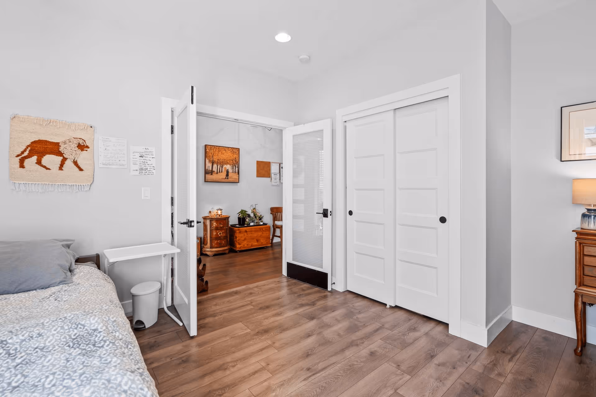 A bright bedroom with a bed on the left side covered with a patterned blanket and a gray pillow. On the wall above the bed is a woven wall hanging depicting a lion. There is a small white table and a trash bin next to the bed. The room has wooden flooring and white walls. A door with glass panels is open, leading to another room with wooden furniture and artwork on the walls. To the right of the door is a white closet with sliding doors. A wooden nightstand with a lamp is partially visible on the right side.