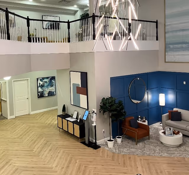 Interior view of a senior living facility lobby or common area with a modern design. The space features a two-story ceiling with a large geometric chandelier hanging down. There is a seating area with a gray sofa, an orange armchair, a round white coffee table, and a tall floor lamp against a blue paneled accent wall with a round mirror. A potted plant is placed near the seating area. On the opposite wall, there is a console table with decorative items and framed pictures, along with abstract artwork hanging above it. The flooring is light wood arranged in a herringbone pattern, and a hallway with a closed white door is visible in the background.