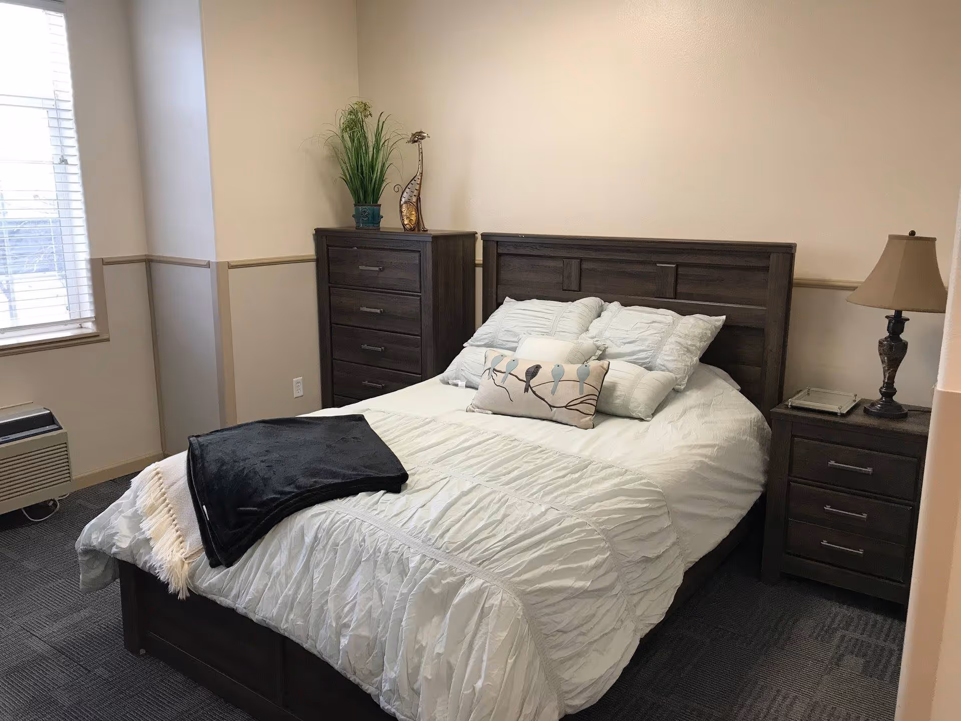 A neatly made bed with white bedding and multiple pillows in a bedroom. The room has a dark wooden headboard, matching nightstand with a lamp, and a tall dresser with a plant and decorative item on top. There is a window with blinds and a wall-mounted air conditioning unit below it.