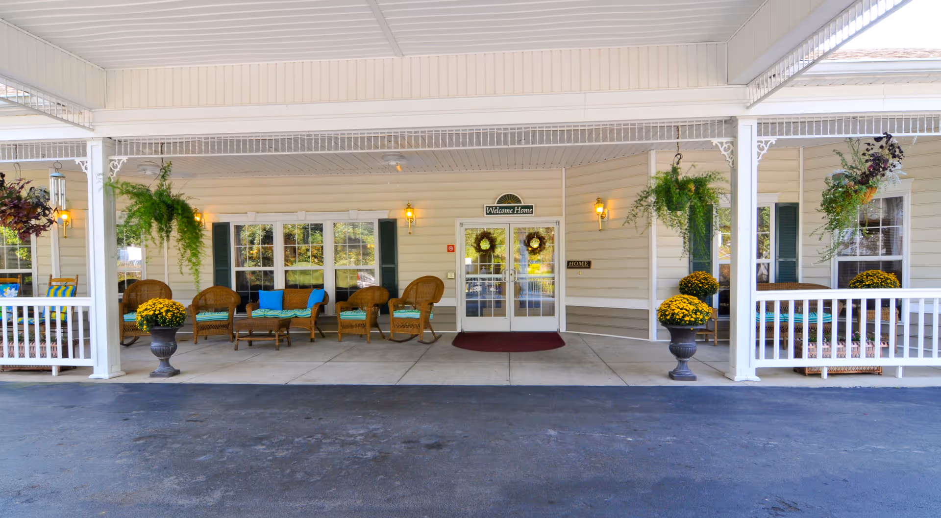 Covered front entrance of a senior living facility with wicker chairs, potted flowers, hanging ferns and double glass doors under a porch.