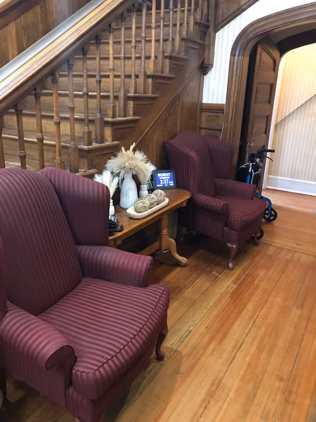 Two maroon striped armchairs flank a wooden side table under a carved wooden staircase in a senior living facility foyer, with a walker visible in the doorway.