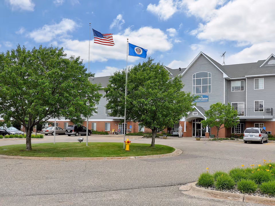 Front exterior of a three-story senior living building showing two flagpoles, trees, parked cars, and a circular driveway.