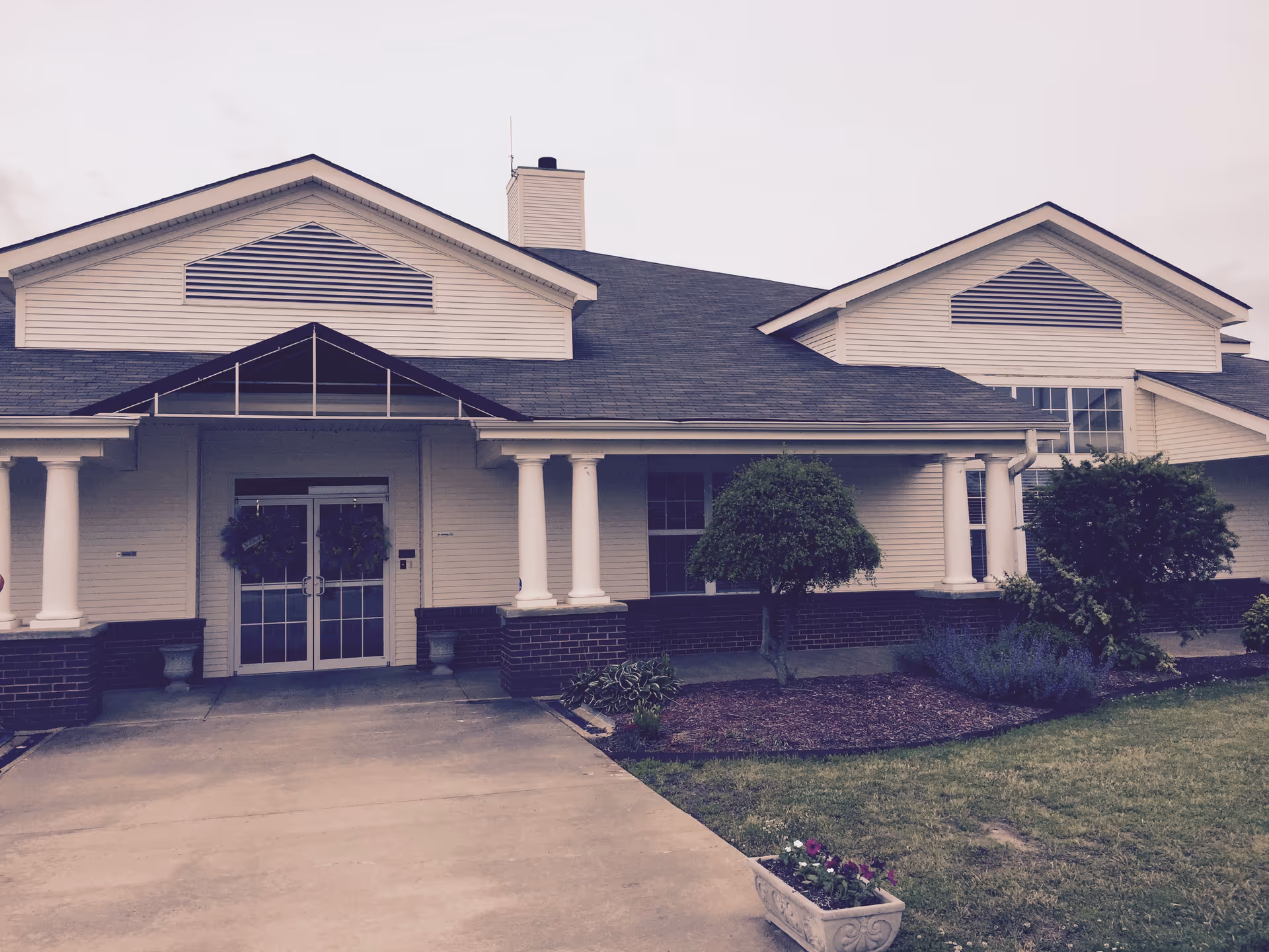 Front exterior view of a single-story senior living facility building with white siding, a dark shingled roof, white columns supporting a covered entrance, glass double doors with wreaths, and landscaped bushes and flowers in front.