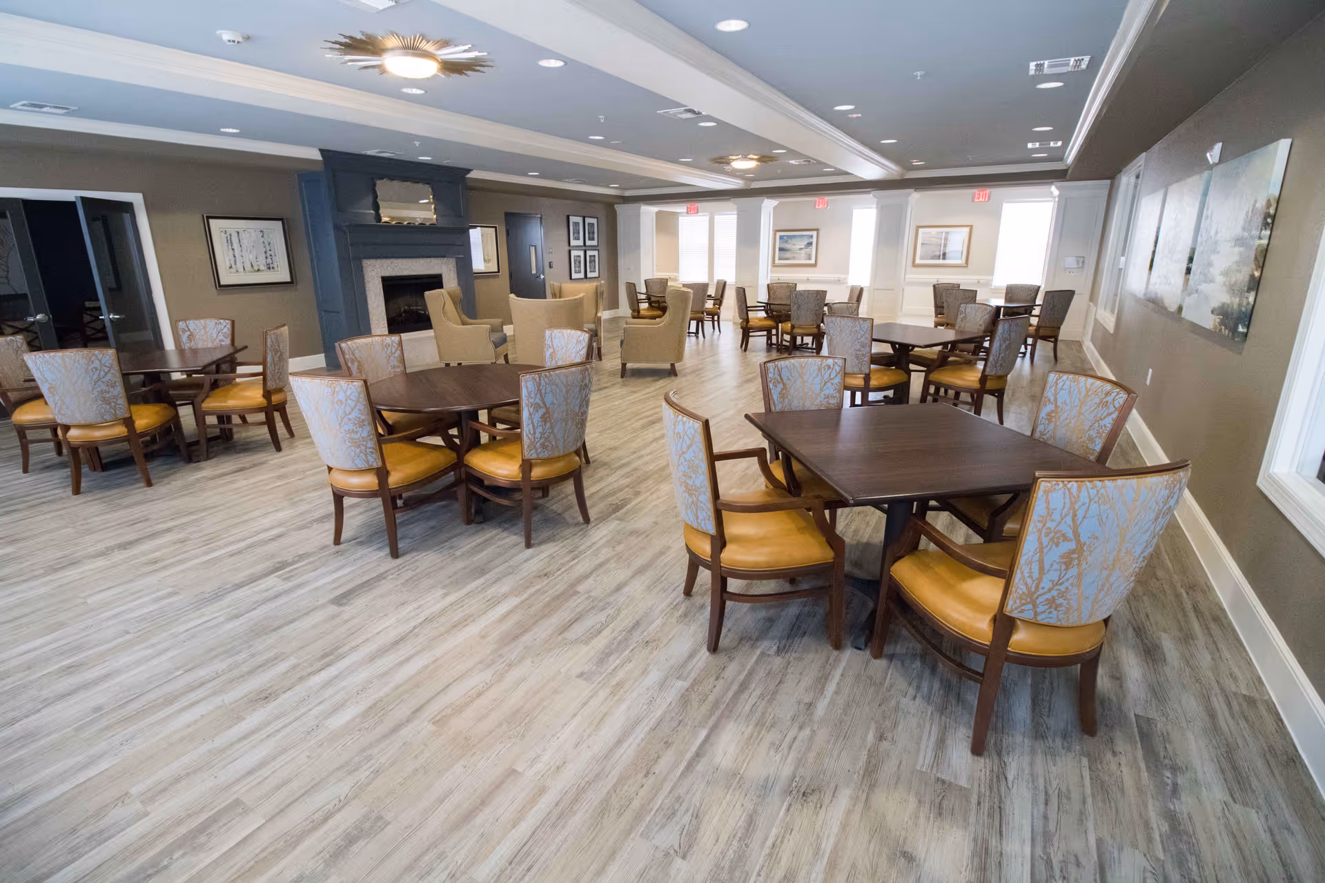 A spacious dining room with multiple wooden tables and cushioned chairs arranged neatly. The room features light wood flooring, neutral-colored walls, large windows letting in natural light, and a fireplace with a mirror above it. Several framed artworks are hung on the walls, and ceiling lights provide additional illumination.