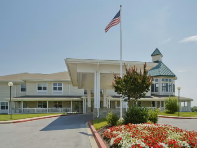 Front exterior view of a senior living facility with a covered entrance, an American flag on a flagpole, and a landscaped flower bed with red and white flowers in the foreground under a clear blue sky.