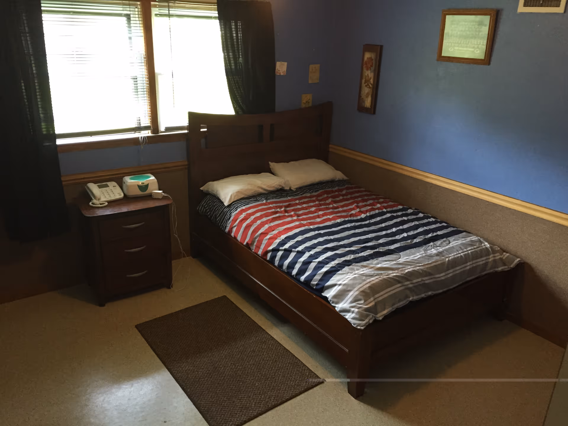 A bedroom in a senior living facility featuring a bed with striped bedding, a bedside table, and a telephones.