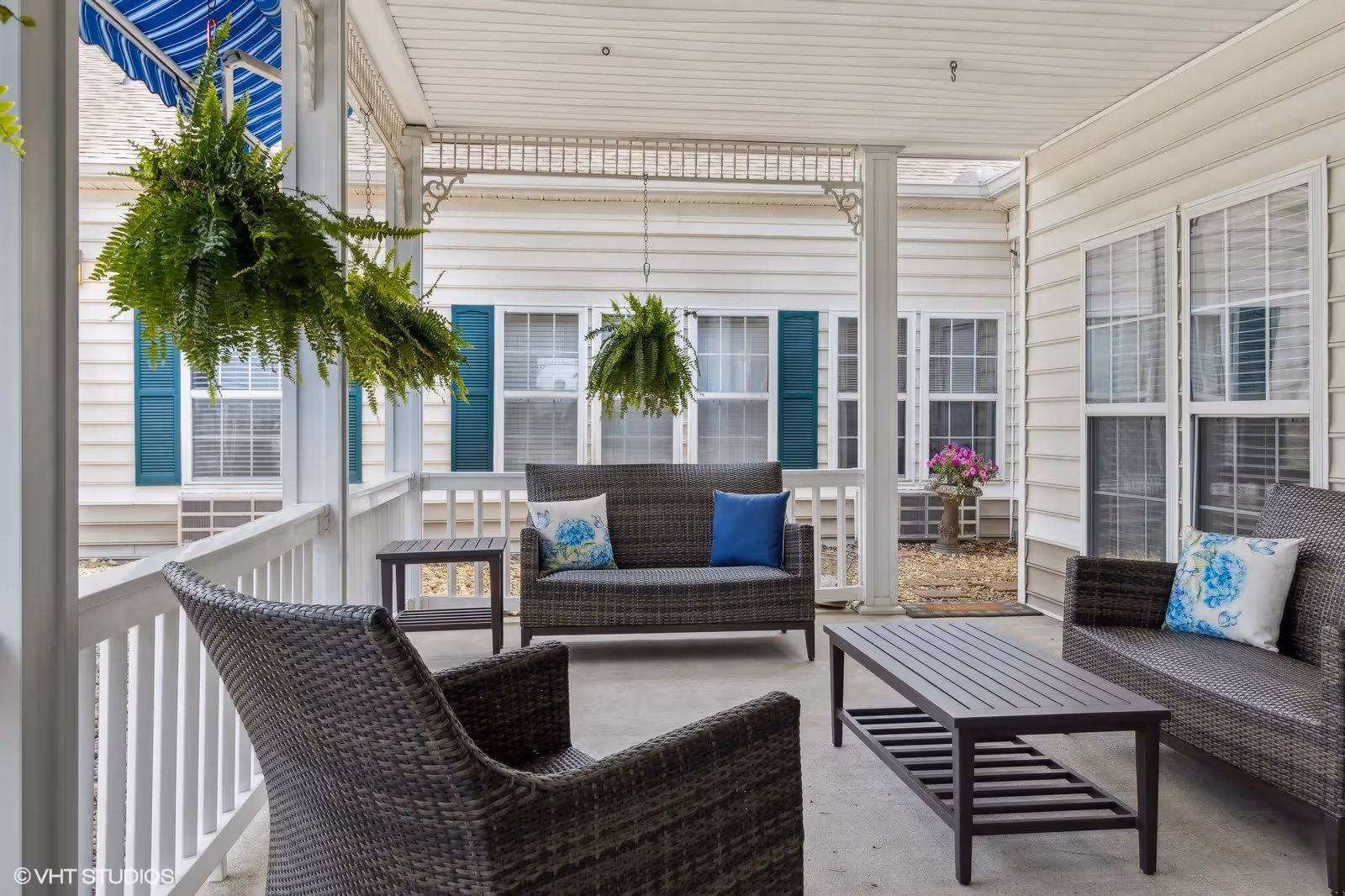 Covered outdoor patio with wicker seating, a coffee table, hanging ferns and decorative pillows in front of a light-sided building.