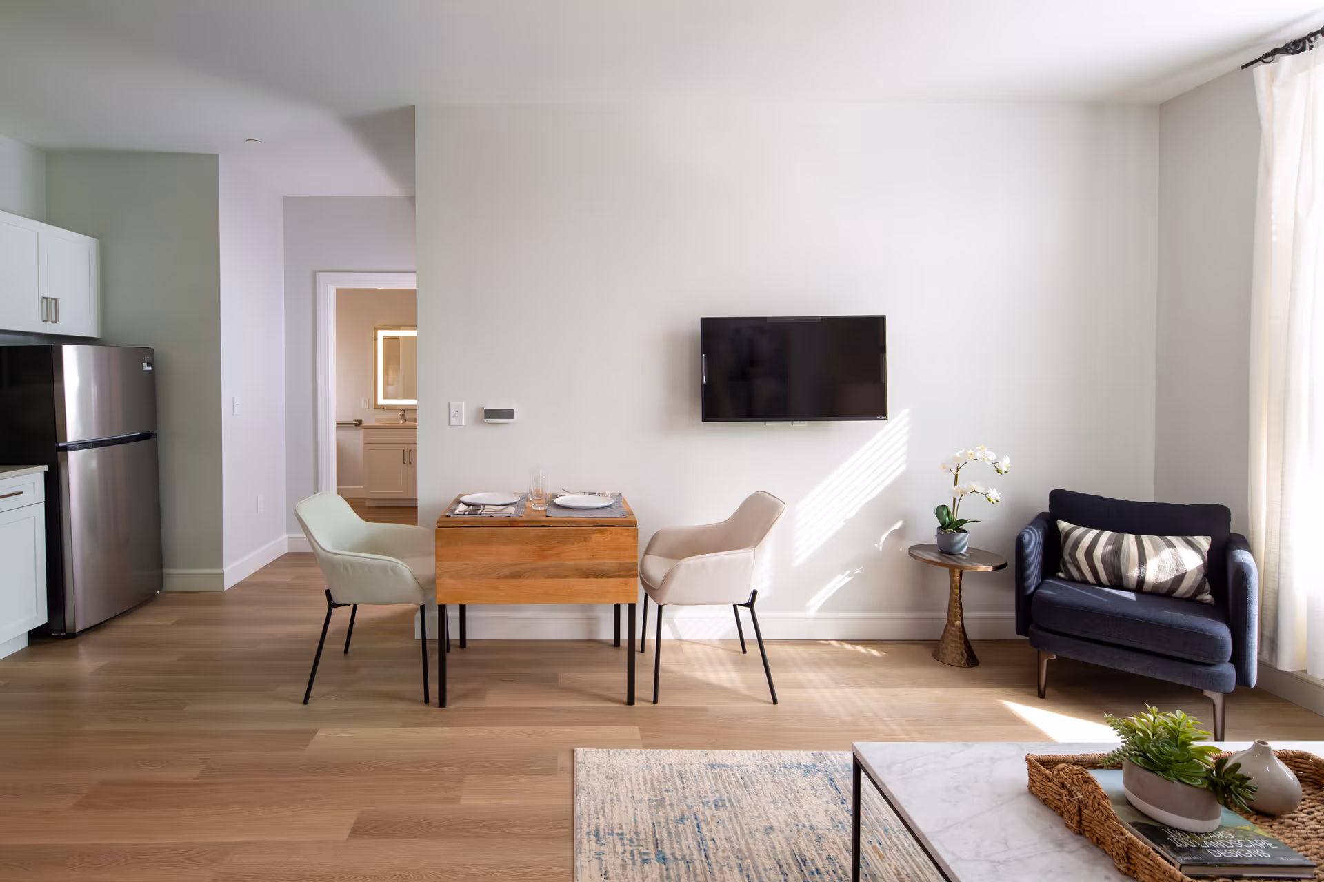 Bright open living area with a small wooden dining table, wall-mounted TV, armchair, and a partial kitchen with refrigerator visible.