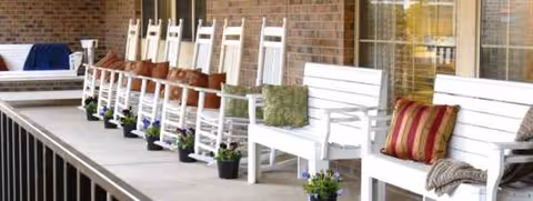 Row of white rocking chairs on a covered porch with potted flowers beside brick wall.