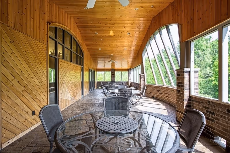 A spacious enclosed patio area with a wooden ceiling and walls, brick lower walls, and large angled windows letting in natural light. The floor is stone-tiled, and there are several round glass tables with metal chairs arranged along the space. Ceiling fans are mounted on the wooden ceiling.
