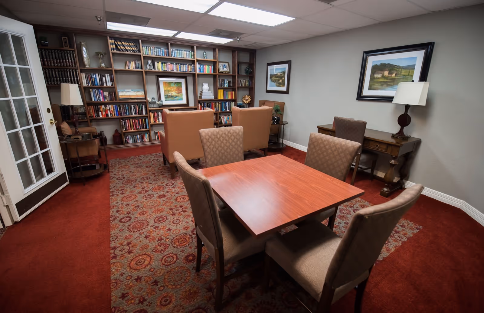 A cozy room with a wooden table surrounded by four upholstered chairs in the foreground. In the background, there are two armchairs facing a large built-in bookshelf filled with books and decorative items. The room has a patterned carpet, framed paintings on the walls, a wooden desk with a lamp, and a glass-paneled door on the left side.