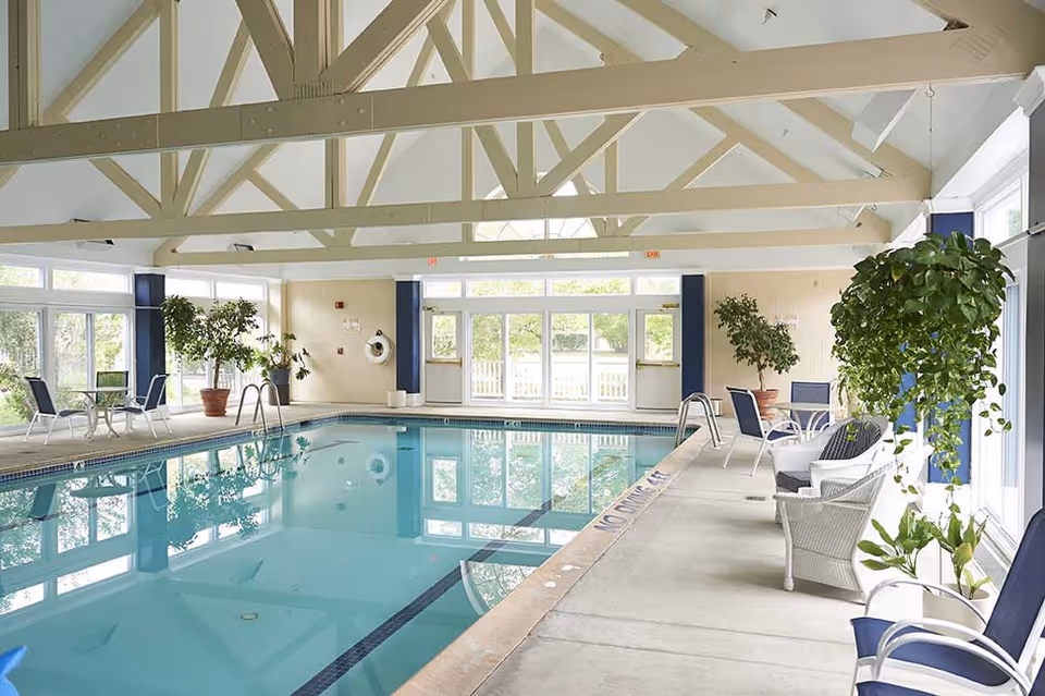 Indoor swimming pool area with clear blue water, surrounded by chairs, tables, and potted plants. The space has large windows allowing natural light to fill the room and a high ceiling with exposed beams.