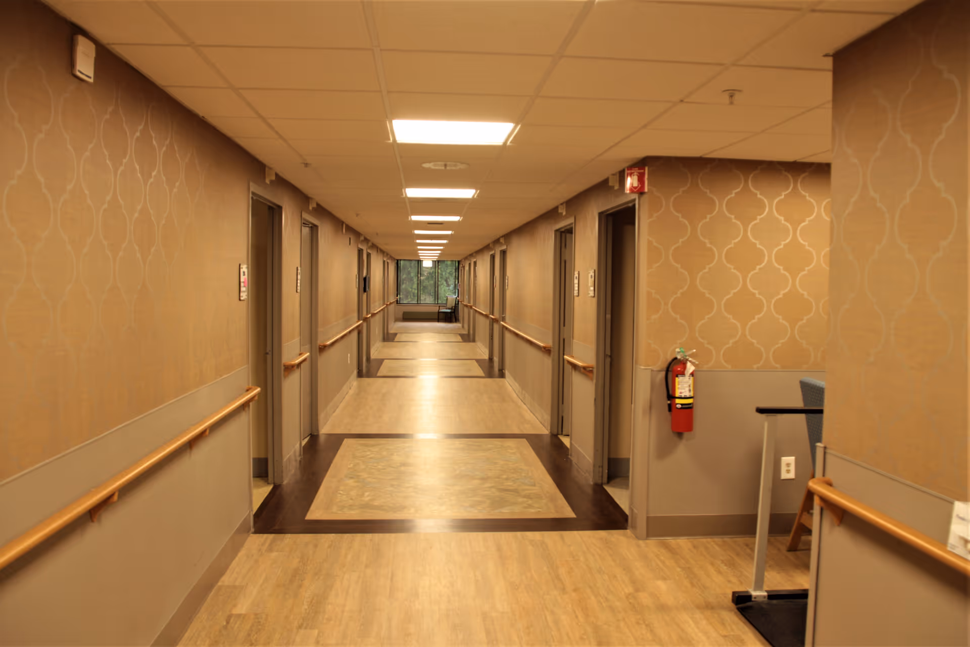 Long, well-lit nursing facility hallway with handrails, doors on both sides, and a fire extinguisher on the wall.