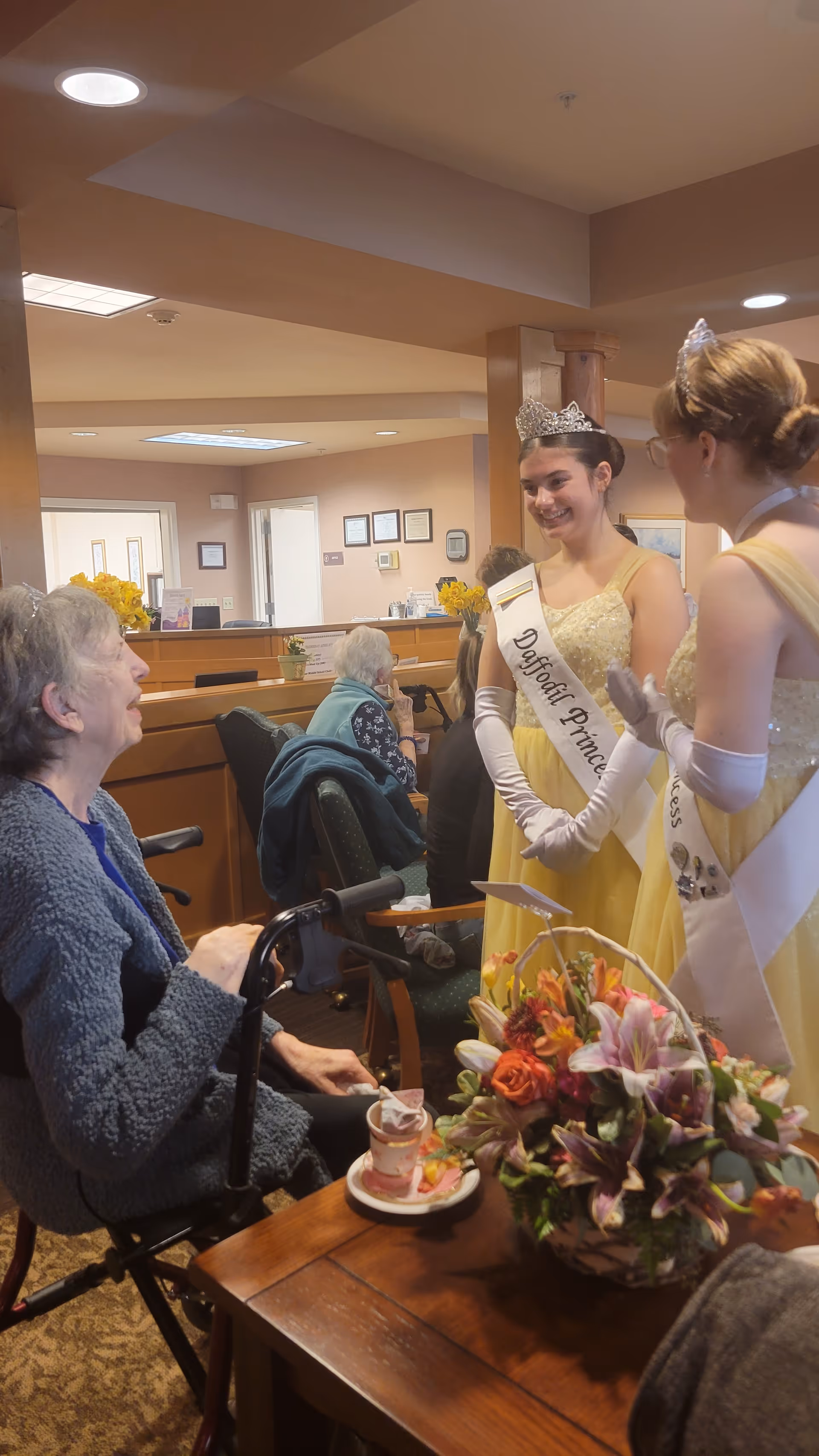 Two young women wearing yellow dresses, white gloves, tiaras, and sashes that read 'Daffodil Princess' are smiling and talking to an elderly woman seated in a wheelchair inside a senior living facility. There is a wooden table with a floral arrangement and a cup in the foreground, and other elderly residents are visible in the background.