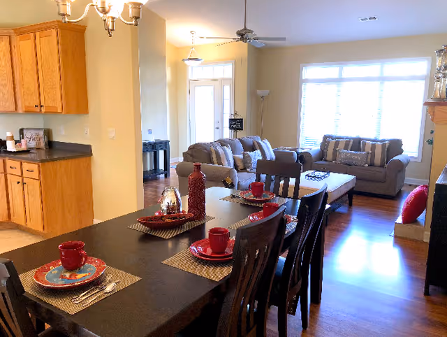 A cozy open-concept living and dining area in a senior living facility. The foreground features a dark wooden dining table set with red plates, cups, and silverware on woven placemats. Behind the dining area is a living room with two beige sofas adorned with striped and solid pillows, a coffee table, and a large window letting in natural light. To the left, there is a kitchen area with wooden cabinets and dark countertops. The room has warm yellow walls and wooden flooring.