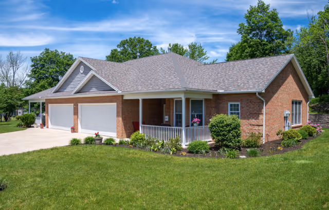 Exterior view of a single-story brick building with a three-car garage, a covered porch with white railings, and well-maintained landscaping including green grass, bushes, and flowers under a blue sky with some clouds.