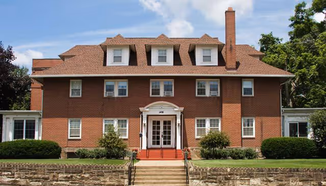 Front exterior view of a large brick building with multiple windows, a central entrance with white double doors, and a red roof with dormer windows. There are bushes and a stone retaining wall in front of the building under a partly cloudy sky.