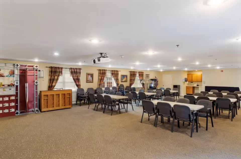A large, well-lit room with multiple rectangular tables and black chairs arranged in rows, suitable for group activities or dining. The room has beige walls, carpeted floor, several windows with plaid curtains, a piano against one wall, framed pictures, and a small kitchenette area in the back corner.