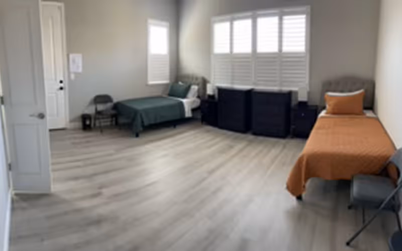 Spacious room with two single beds on opposite sides, dressers under shuttered windows, and folding chairs on light wood flooring.