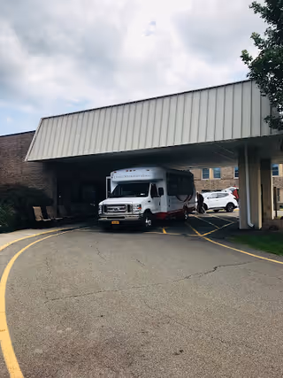 A white shuttle van parked under a covered drop-off at the front entrance of a brick senior living facility.