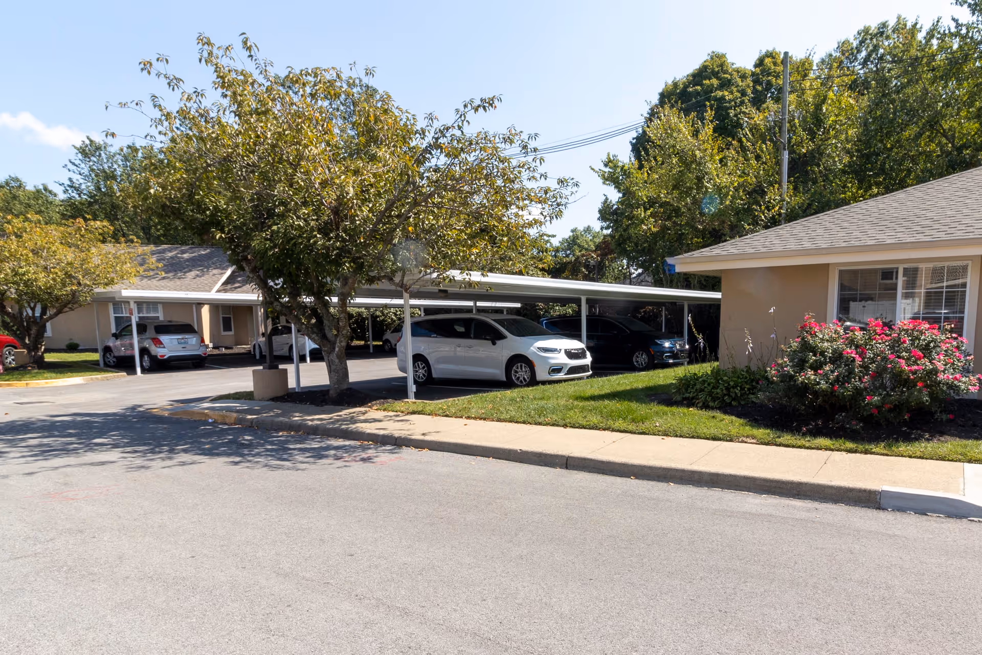 Outdoor view of a senior living facility parking area with several cars parked under a covered carport. There are trees and bushes with flowers near the buildings, and the sky is clear and blue.