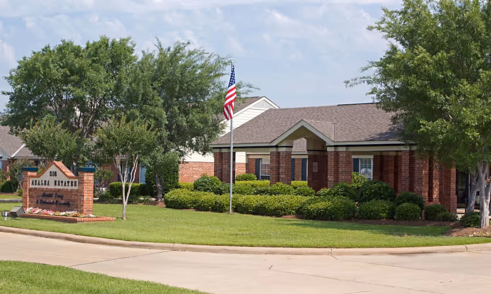 Exterior view of Azalea Estates of New Iberia, showing a single-story brick building with a covered entrance, surrounded by green bushes and trees, with an American flag on a flagpole in front and a sign displaying the facility name and address.