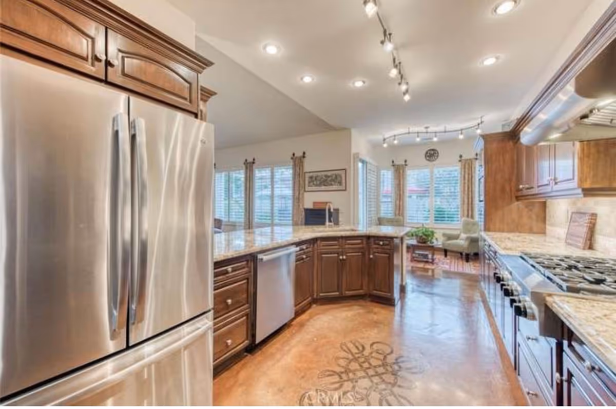 Spacious kitchen with stainless steel refrigerator, granite countertops, wooden cabinets, and a breakfast nook in the background.